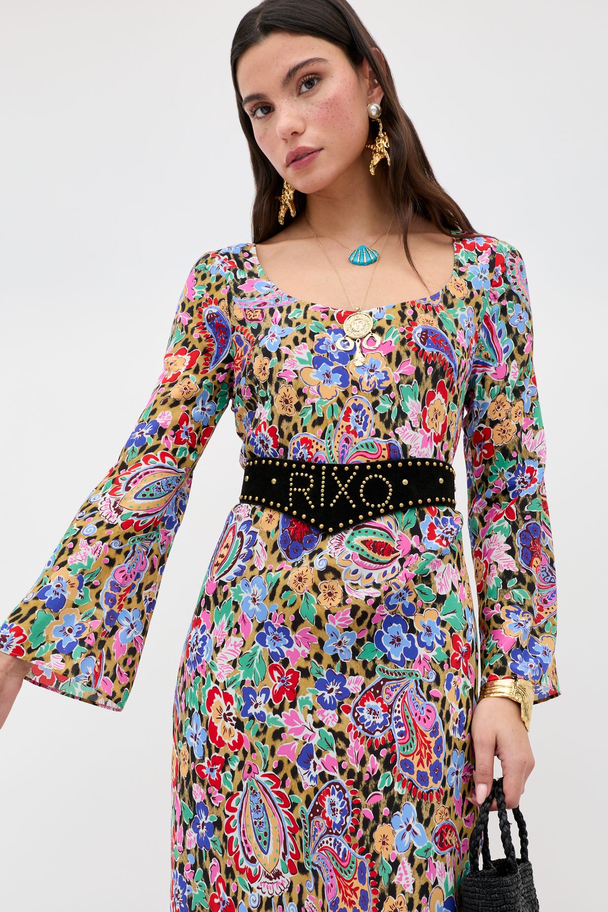 A woman with long dark hair wears the Lorelei - Black, a long-sleeved, multicolored floral and paisley dress cinched at the waist with a black belt. She accessorizes with gold jewelry and carries a black handbag, standing against a plain background.