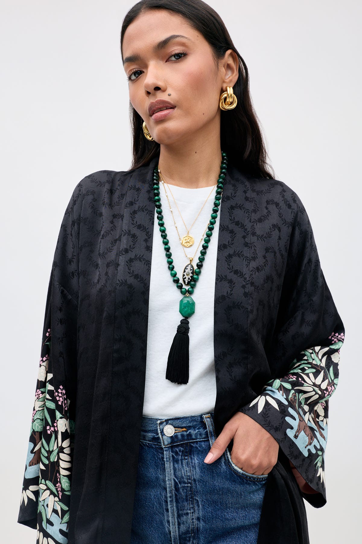A woman wearing the Cally - Green kimono, paired with a white shirt, blue jeans, gold earrings, layered necklaces, and a long beaded tassel necklace stands with one hand in her pocket, posing confidently.