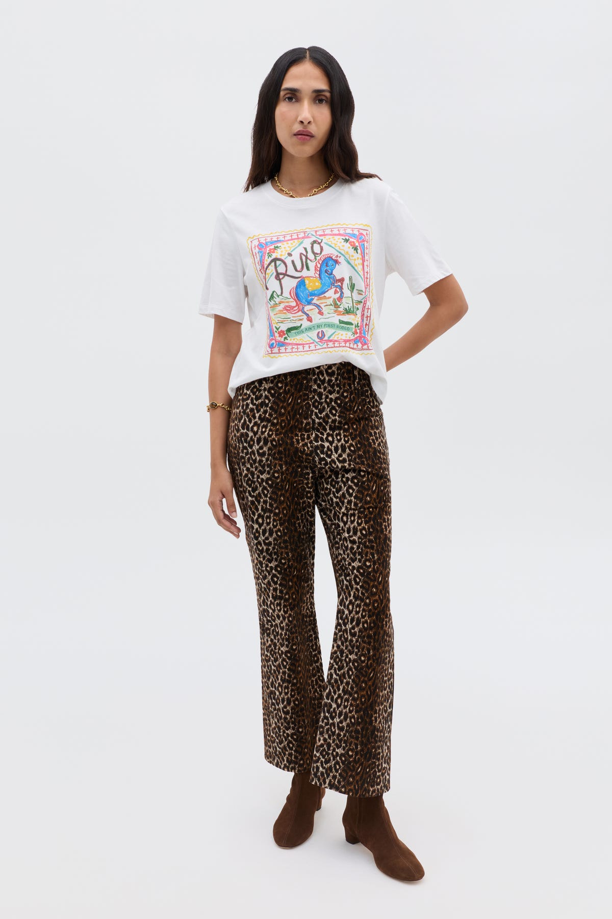A woman stands against a plain background wearing a white printed t-shirt with colorful graphics, Olwen - Lyra Leopard Black trousers, brown ankle boots, and a gold bracelet.