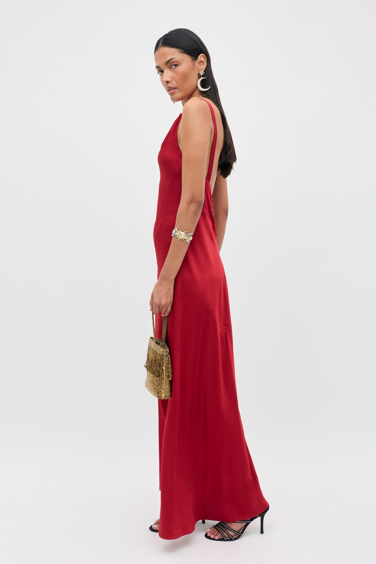 Wearing the Adelina - Red dress with thin straps, she holds a beaded gold handbag and accessorizes with hoop earrings, a bracelet, and black heeled sandals, standing against a plain light background.