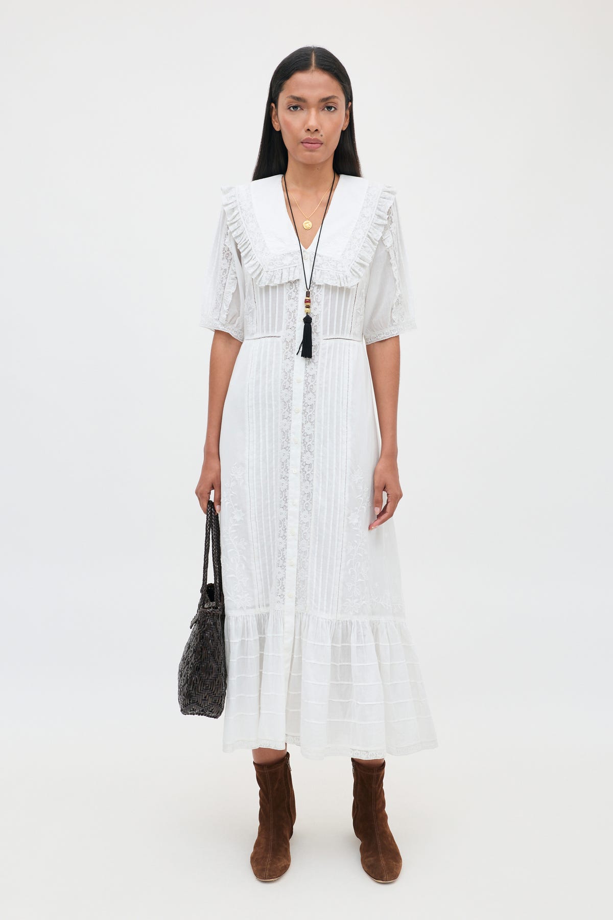 A woman stands against a white background wearing the Edina - Pansy Embroidery White dress, featuring delicate lace embroidery details, paired with brown boots, a black tasseled necklace, and holding a black handbag at her side.