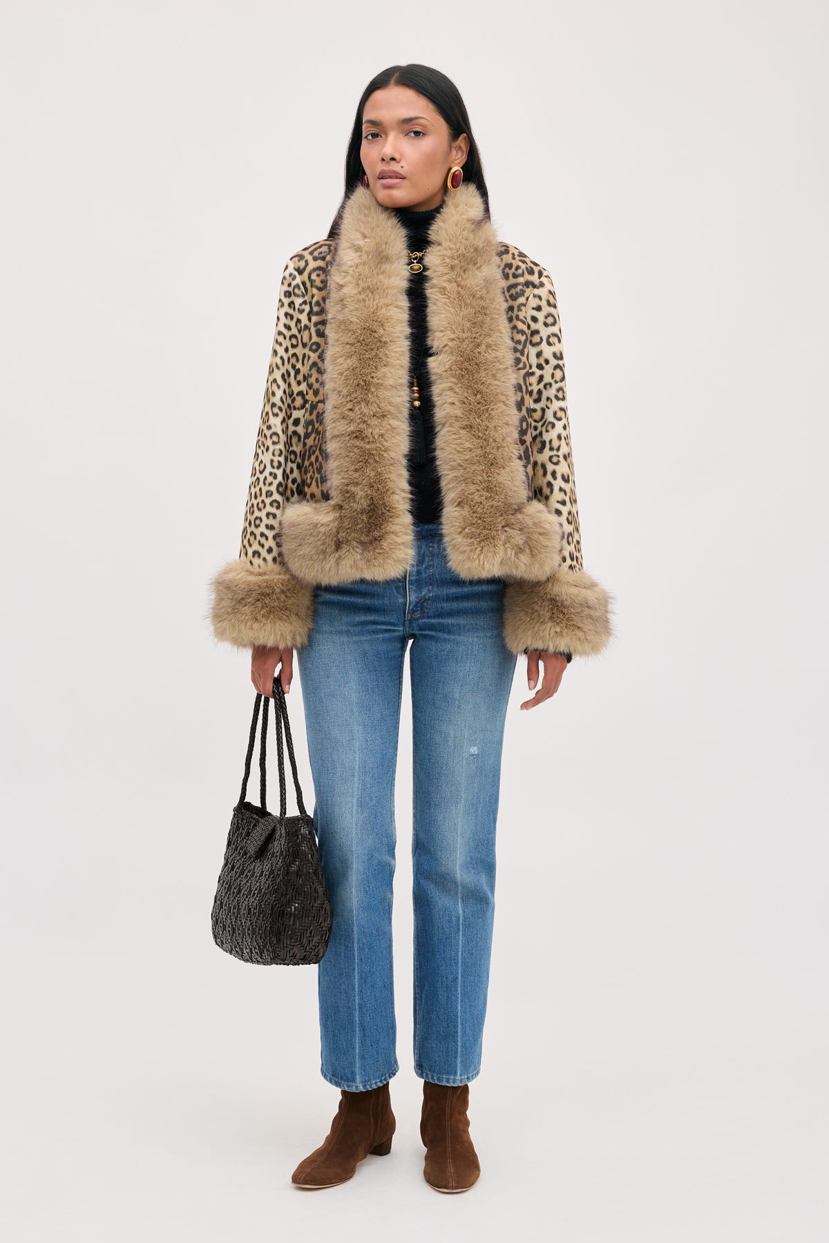 A model wearing a Jorgia - Savannah Leopard Brown jacket with faux fur trim, a black turtleneck, blue jeans, and brown suede ankle boots, carrying a dark woven handbag.