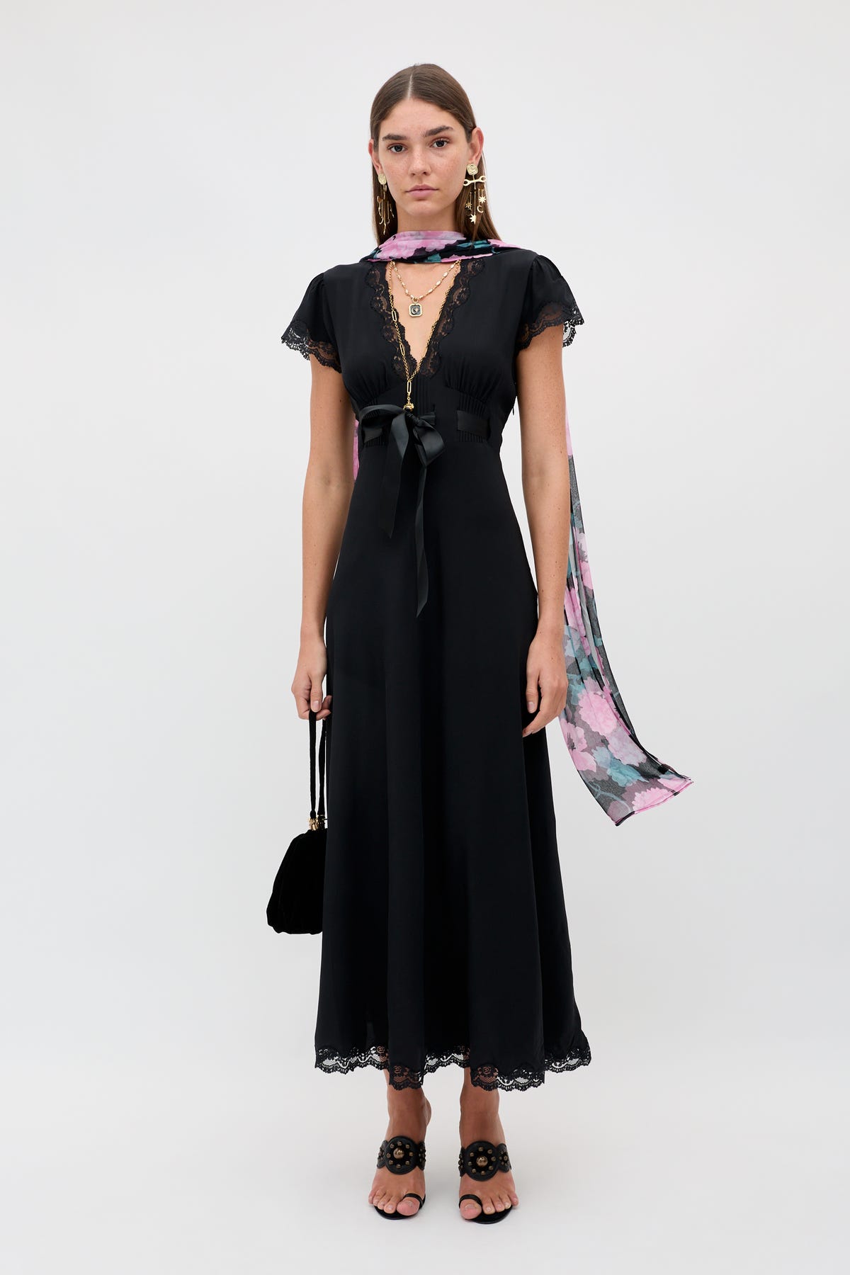 A woman stands against a plain white background wearing the Marlice - Black: a black, short-sleeved, ankle-length dress with lace trim. She accessorizes her look with statement earrings, a pink floral scarf tied in a delicate satin bow, black heeled sandals, and carries a coordinating black handbag.
