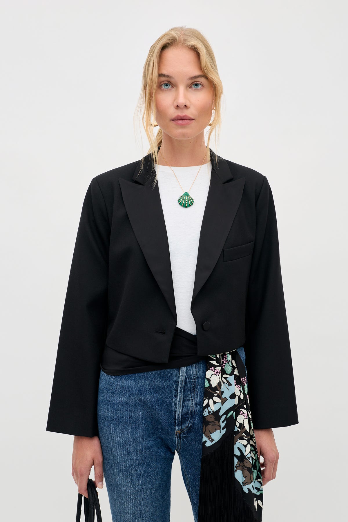 Woman with blonde hair wearing a black blazer, white top, and blue jeans, holding the Meria - Black bag. She has a green pendant necklace and a floral-print scarf draped over her waist, standing against a plain background.