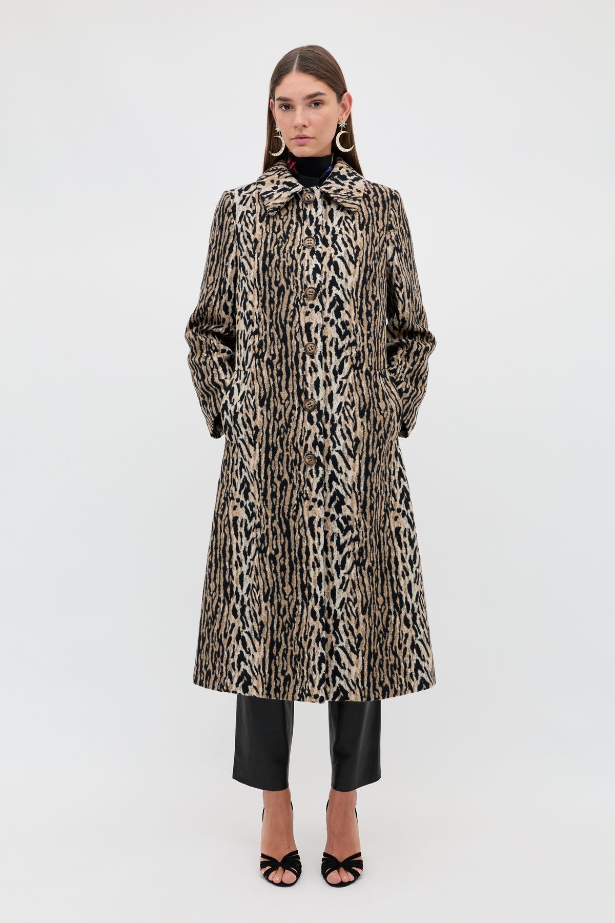 A woman stands against a plain background wearing the Milly - Bohemia Leopard mid-length coat, styled with a black turtleneck, black pants, black open-toe heels, and large hoop earrings. Her hands are in her pockets and her hair is straight.