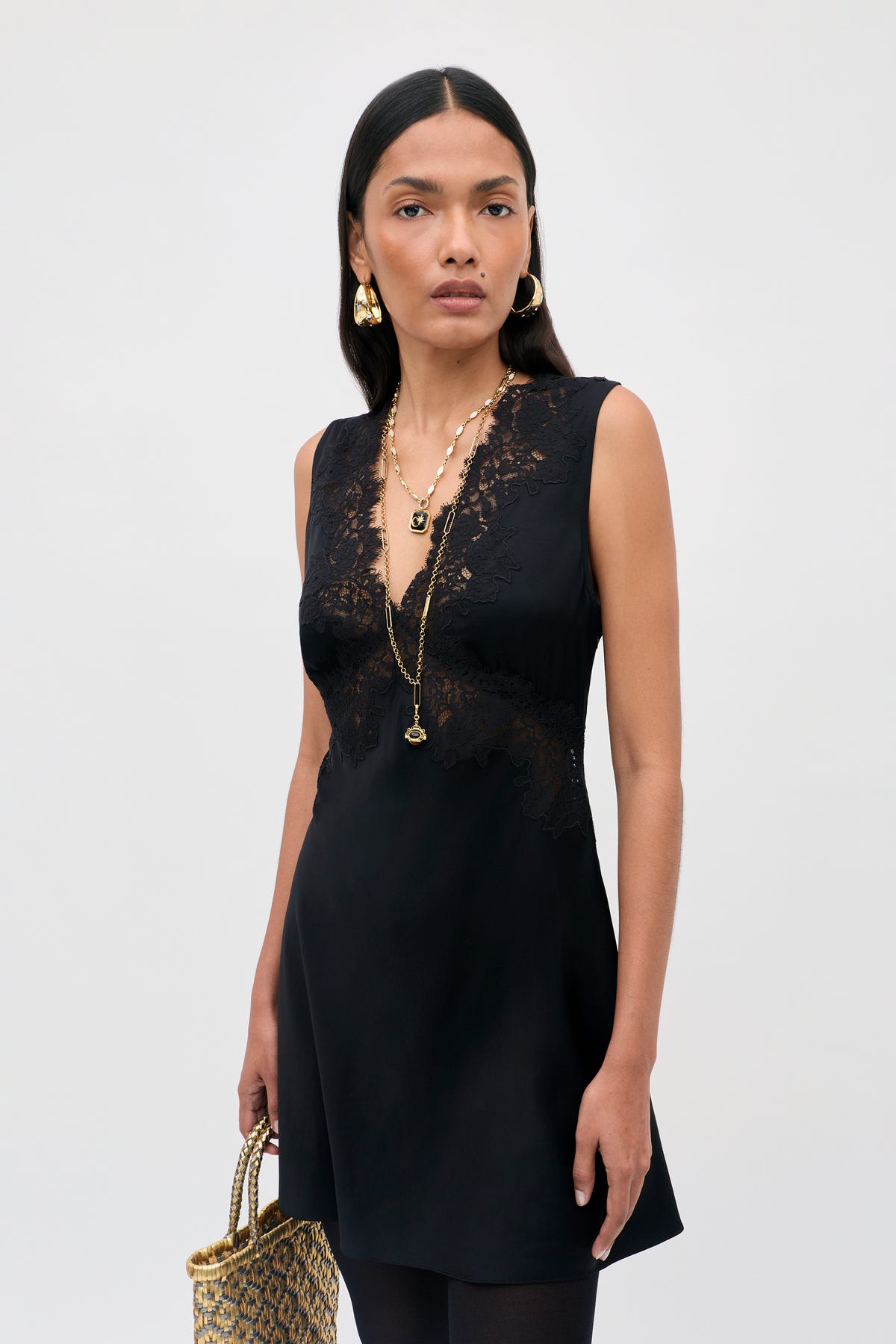 A woman wearing the Rhegan - Black sleeveless dress with lace details, layered gold necklaces, gold earrings, and holding a woven handbag stands against a plain light background.
