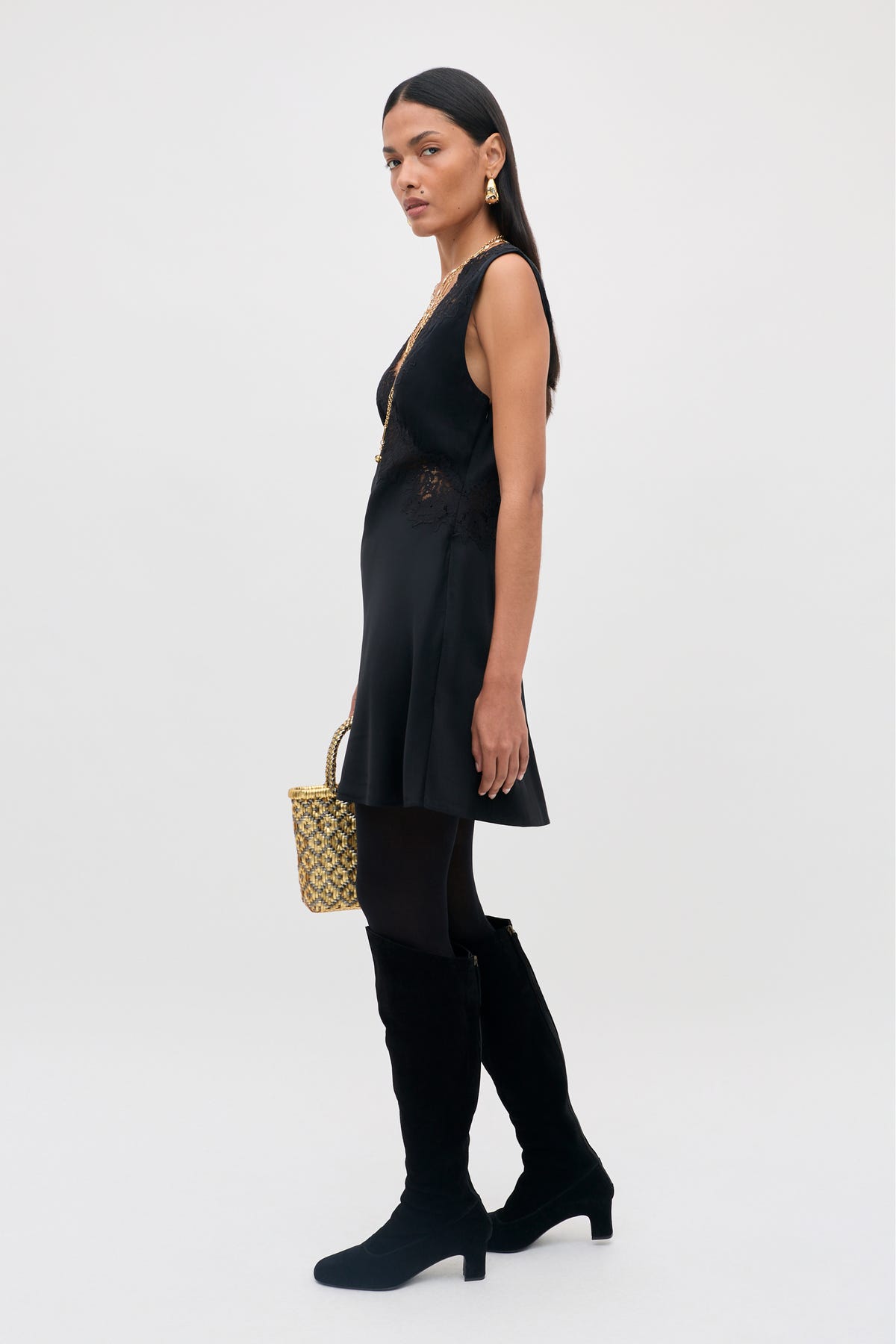 Dressed in the Rhegan - Black, a sleeveless black dress, with black tights and knee-high black boots, a woman stands holding a small patterned handbag. Her long dark hair frames her face, adorned with gold earrings, as she poses with a neutral expression against a light backdrop.