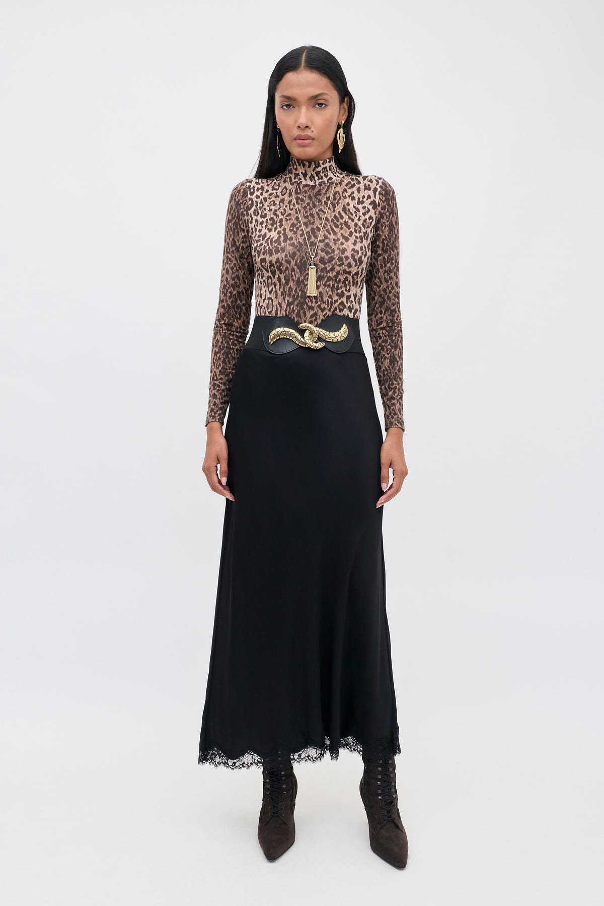A woman stands facing forward against a plain white background, wearing the Stassie - Black matte satin skirt with wavy lace trim, paired with a long-sleeve leopard print top, a black belt featuring a gold snake buckle, brown boots, and gold earrings.