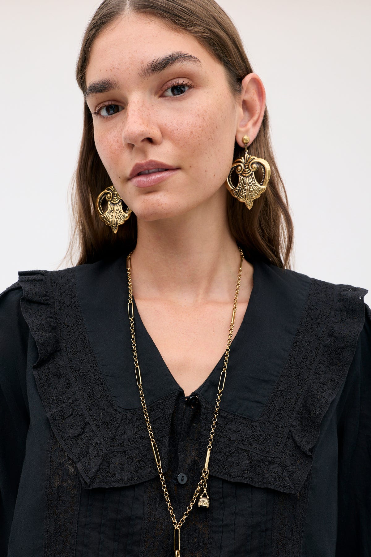 A person wearing Julieta Earrings Gold, featuring an ornate shield-like design, paired with a long gold link chain necklace.
