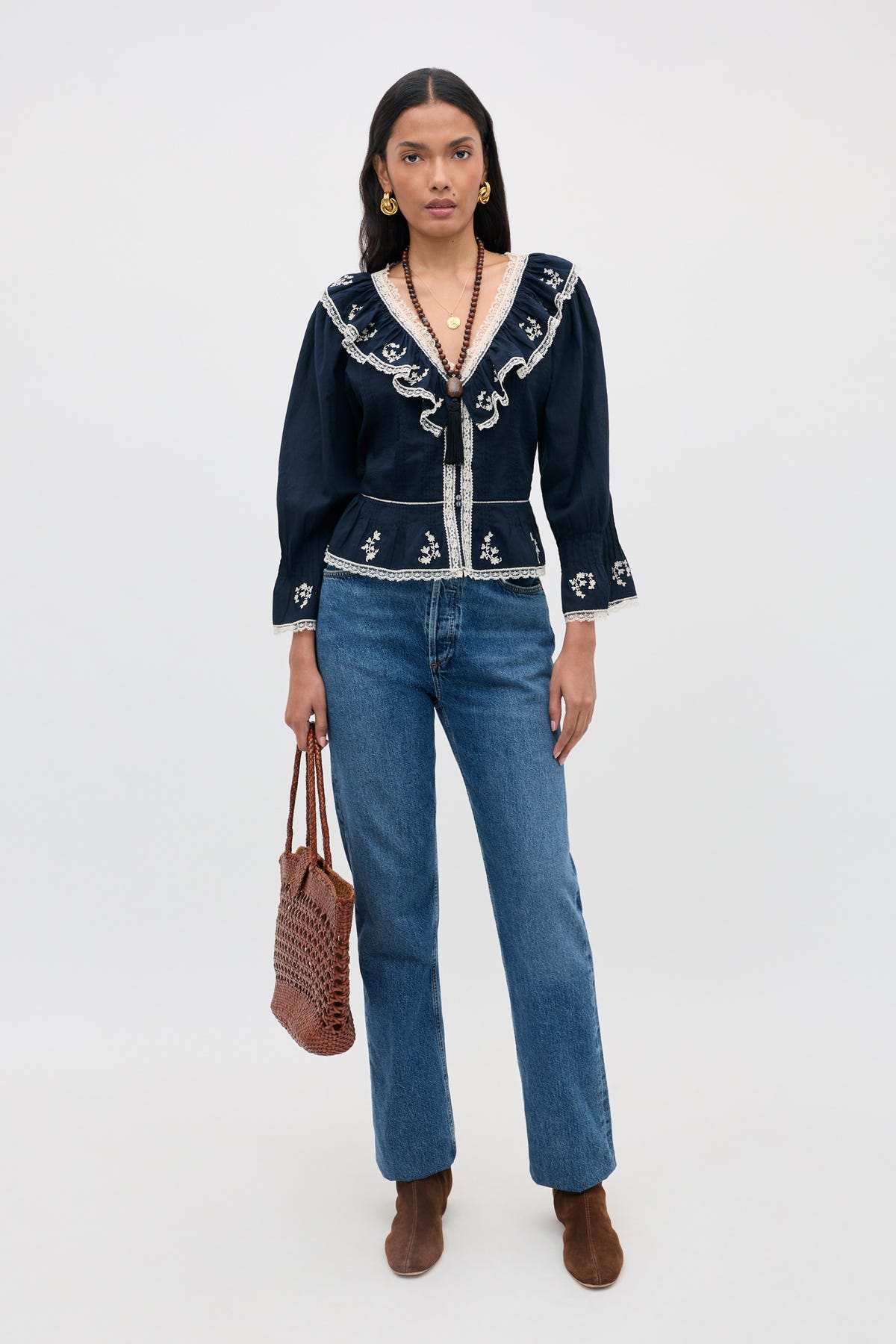 A woman stands against a plain light background wearing the solene top flower trail embroidery navy—a navy ruffled V-neck cotton blouse with white embroidery—paired with blue jeans, brown boots, gold jewelry, and holding a woven brown bag.