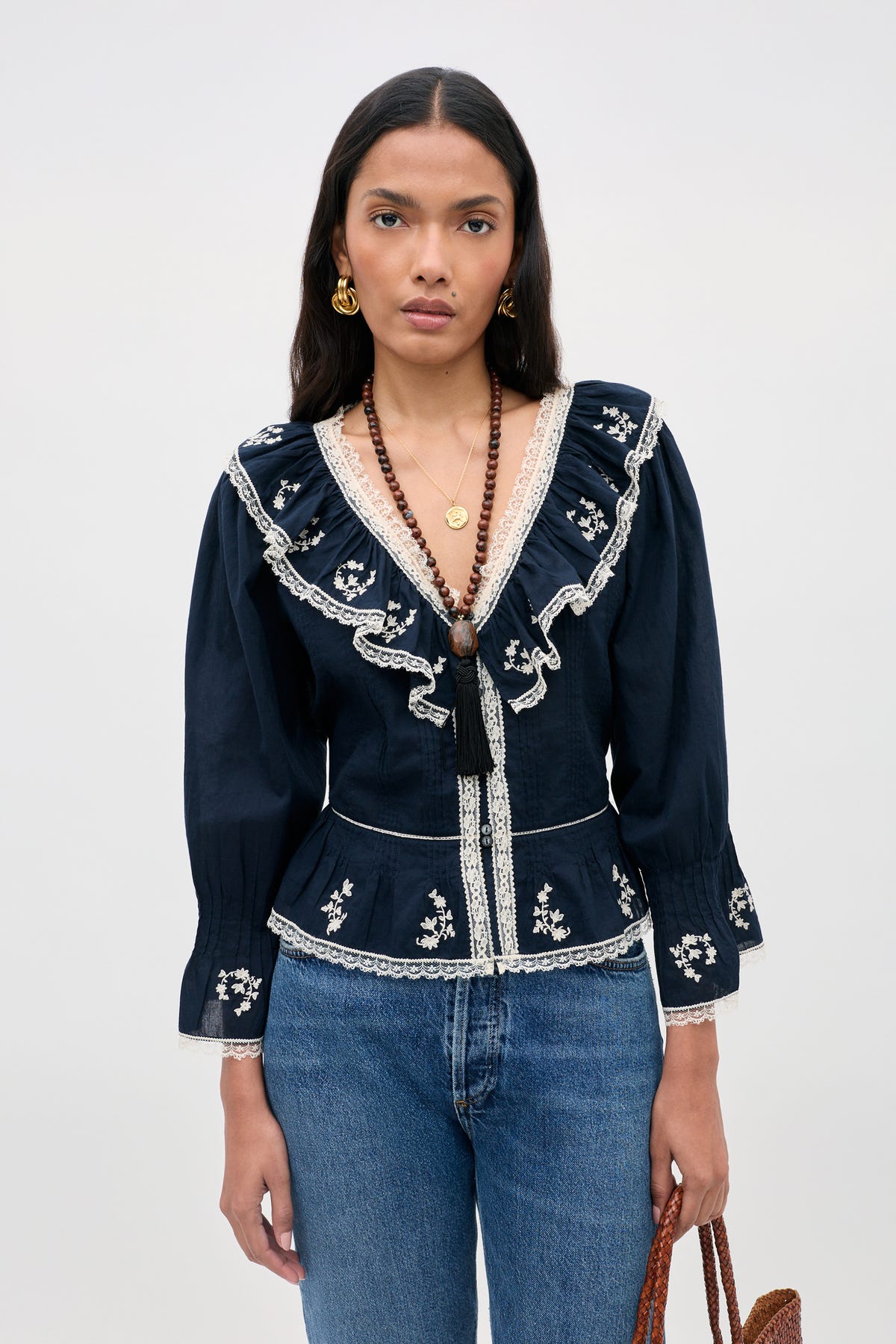 A woman is wearing the Solene Top Flower Trail Embroidery Navy—a navy cotton blouse adorned with white floral embroidery, delicate lace trim, and long sleeves—paired with blue jeans. The vintage-inspired top features a ruffled V-neck that lends an elegant touch to her look. She has long dark hair, gold earrings, layered necklaces, and carries a brown bag while standing against a plain light background.