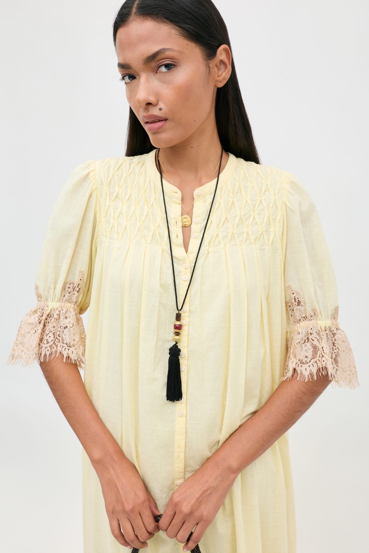 A person wearing the Lorela - Lemon pale yellow button-down blouse with gathered detailing and beige lace-trimmed sleeves, accessorized with two layered necklaces.