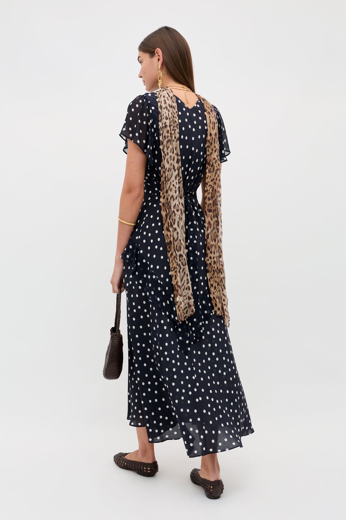 A woman stands facing away, wearing the Evie - Bohemia Spot Deep Navy dress with white polka dots, a leopard print scarf down her back, brown woven flats, gold jewelry, and a dark brown handbag.