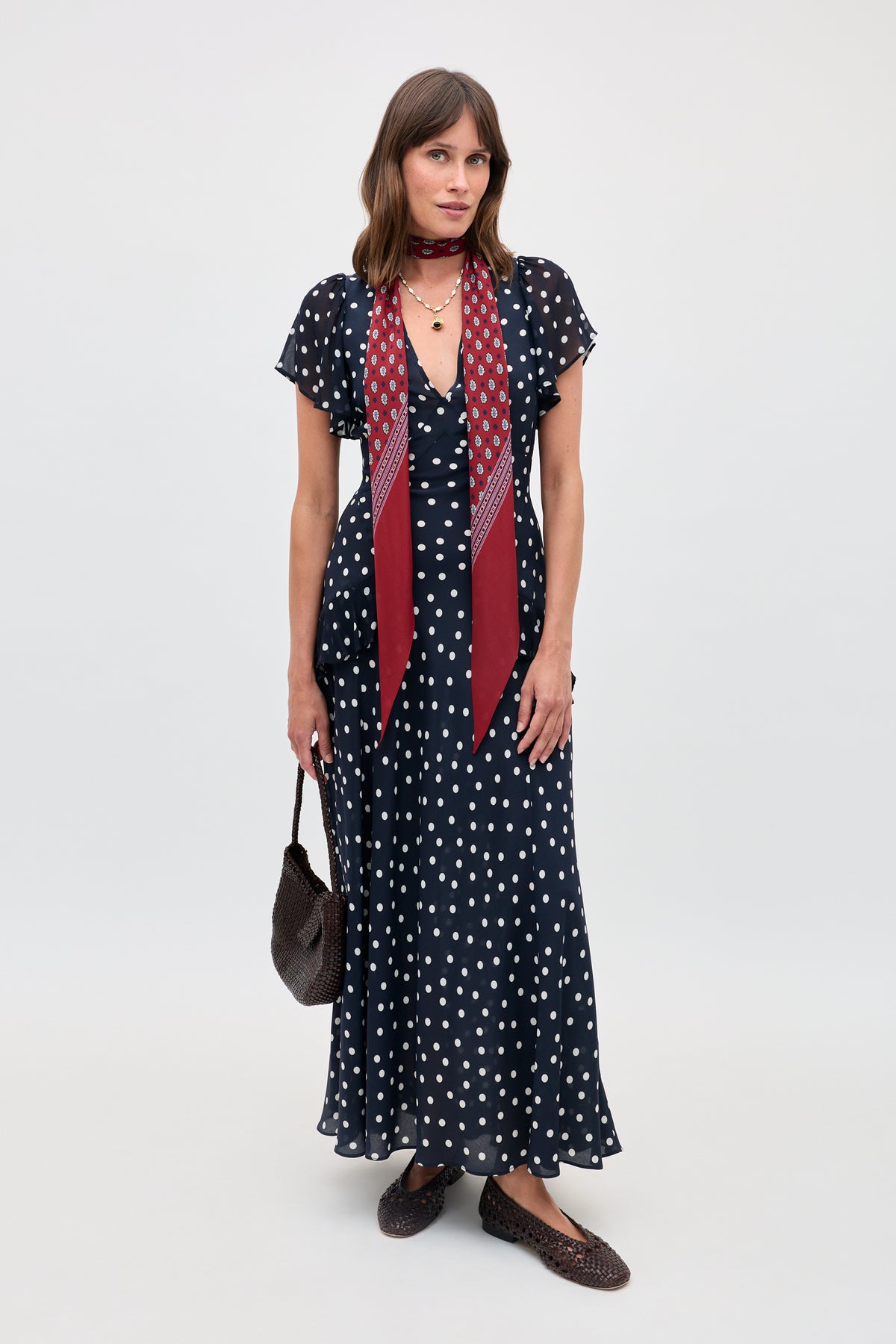 A woman stands against a plain background wearing the Evie - Bohemia Spot Deep Navy dress. She pairs it with a maroon printed scarf, a woven brown bag, and matching woven brown flats.