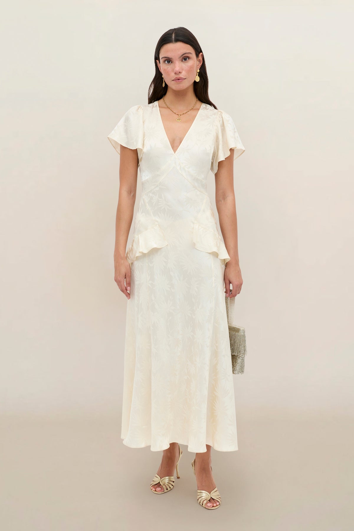 A woman stands against a plain background wearing the Evie - Daisy Jacquard Champagne, a cream-colored, short-sleeved, V-neck midi silk-jacquard dress with a subtle print, styled with strappy heels and a small handbag.