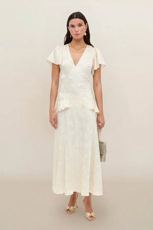 A woman stands against a plain background wearing the Evie - Daisy Jacquard Champagne, a cream-colored, short-sleeved, V-neck midi silk-jacquard dress with a subtle print, styled with strappy heels and a small handbag.