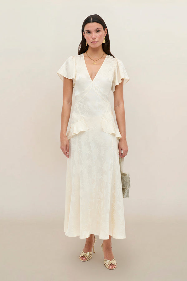 A woman stands against a plain background wearing the Evie - Daisy Jacquard Champagne, a cream-colored, short-sleeved, V-neck midi silk-jacquard dress with a subtle print, styled with strappy heels and a small handbag.