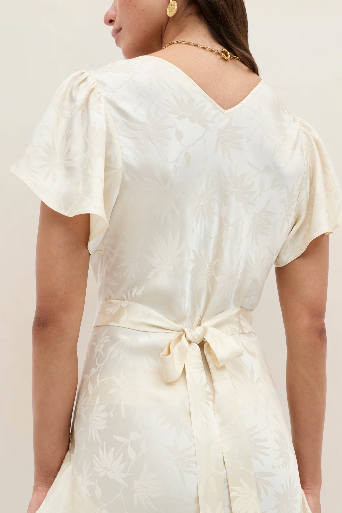 A woman is shown from the back wearing the Evie - Daisy Jacquard Champagne dress, a cream-hued silk-jacquard piece featuring a delicate daisy pattern, flattering ruffles, flutter sleeves, and a waist tie. She has long hair and accessorizes with gold hoop earrings and a gold necklace.