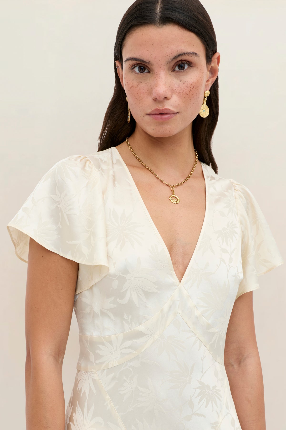 A woman with straight dark hair wears the Evie - Daisy Jacquard Champagne dress, its cream silk-jacquard fabric featuring flattering ruffles at the flutter sleeves. She accessorizes with gold earrings and a matching necklace, standing against a neutral background and looking directly at the camera.