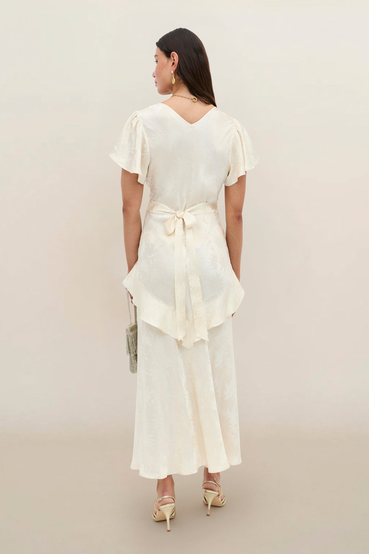 A woman stands with her back to the camera, wearing the Evie - Daisy Jacquard Champagne dress—a light cream, ankle-length silk-jacquard design featuring short sleeves and a waist tie. She holds a small bag and wears light-colored heels against a neutral background. Delicate, flattering ruffles lend an elegant touch to this timeless look.