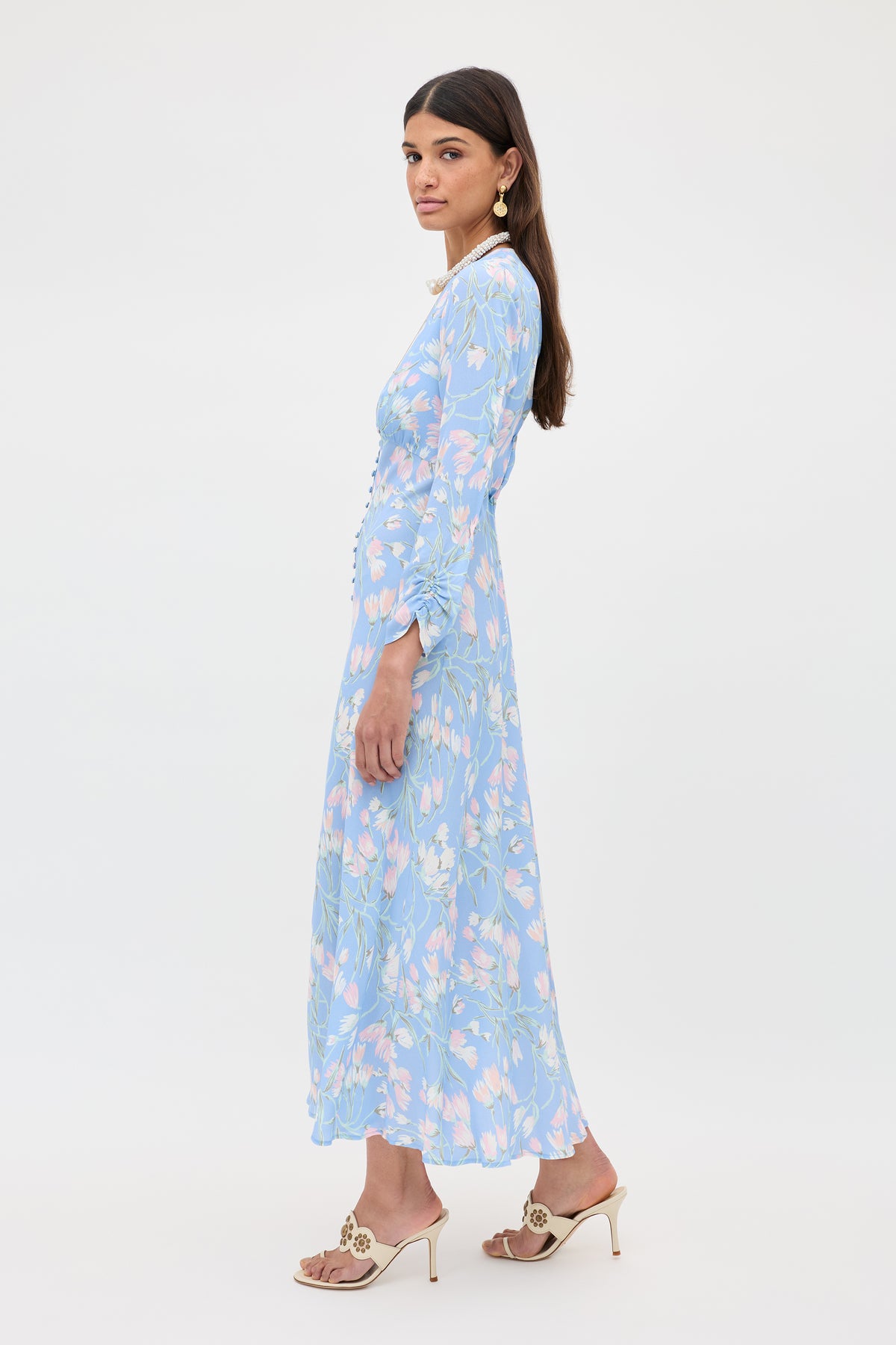 A woman with long brown hair models the Katie - Watercolour Floral Blue dress, a long, light blue floral piece with long sleeves. She wears beige heeled sandals, statement earrings, and a necklace against a plain white background.