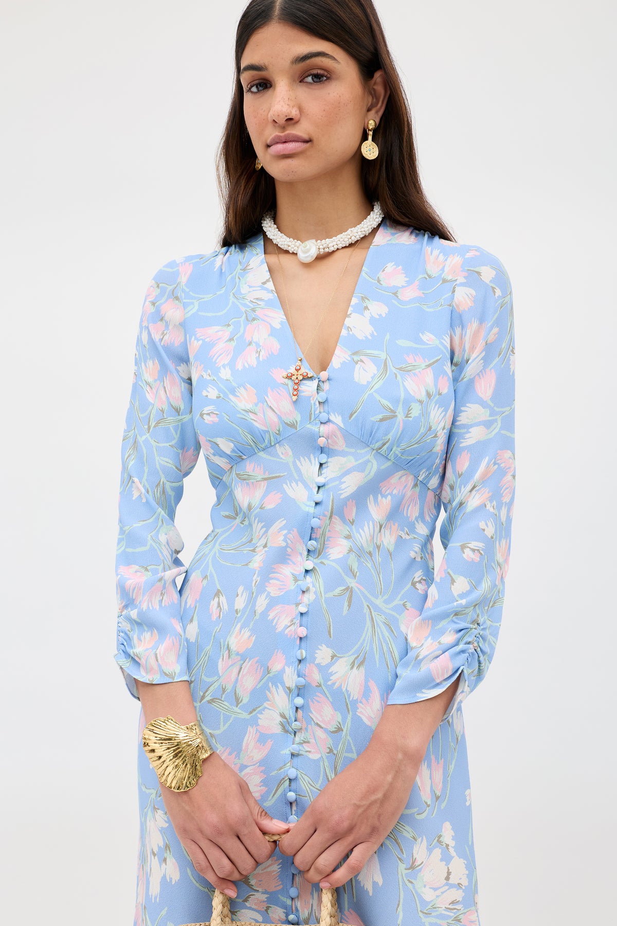 A woman wears the Katie - Watercolour Floral Blue dress, featuring long sleeves and covered buttons. She accessorizes with a gold bracelet, pearl necklace, and gold earrings against a plain light background.