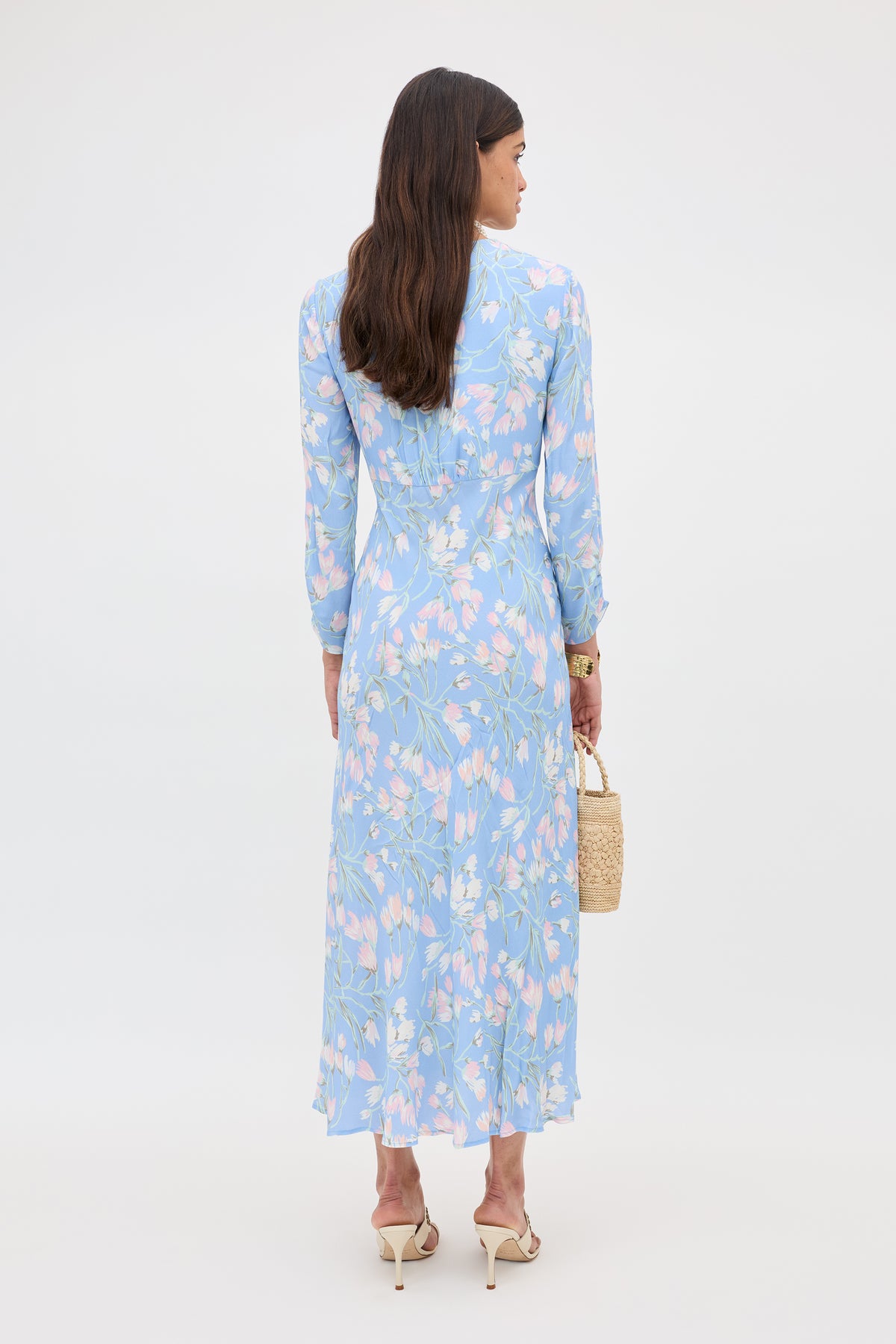 A woman with long brown hair faces away, wearing the Katie - Watercolour Floral Blue midi dress with long sleeves and beige heels. She holds a small woven bag, standing against a plain white background.