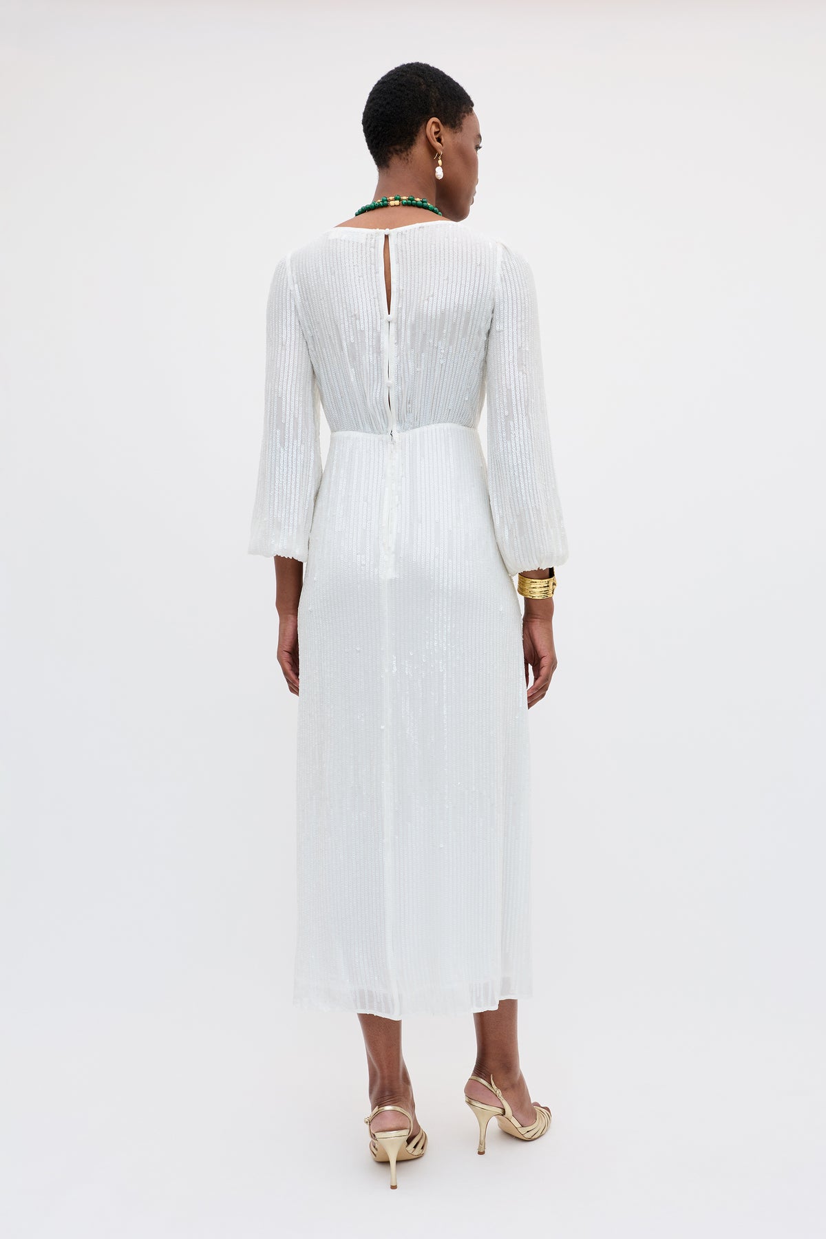 A woman facing away wears the Coco - Ivory dress, an ivory sequin gown with vertical pleats, a keyhole back, and long sleeves. She pairs it with gold sandals, a green necklace, and gold bracelets against a plain white background.