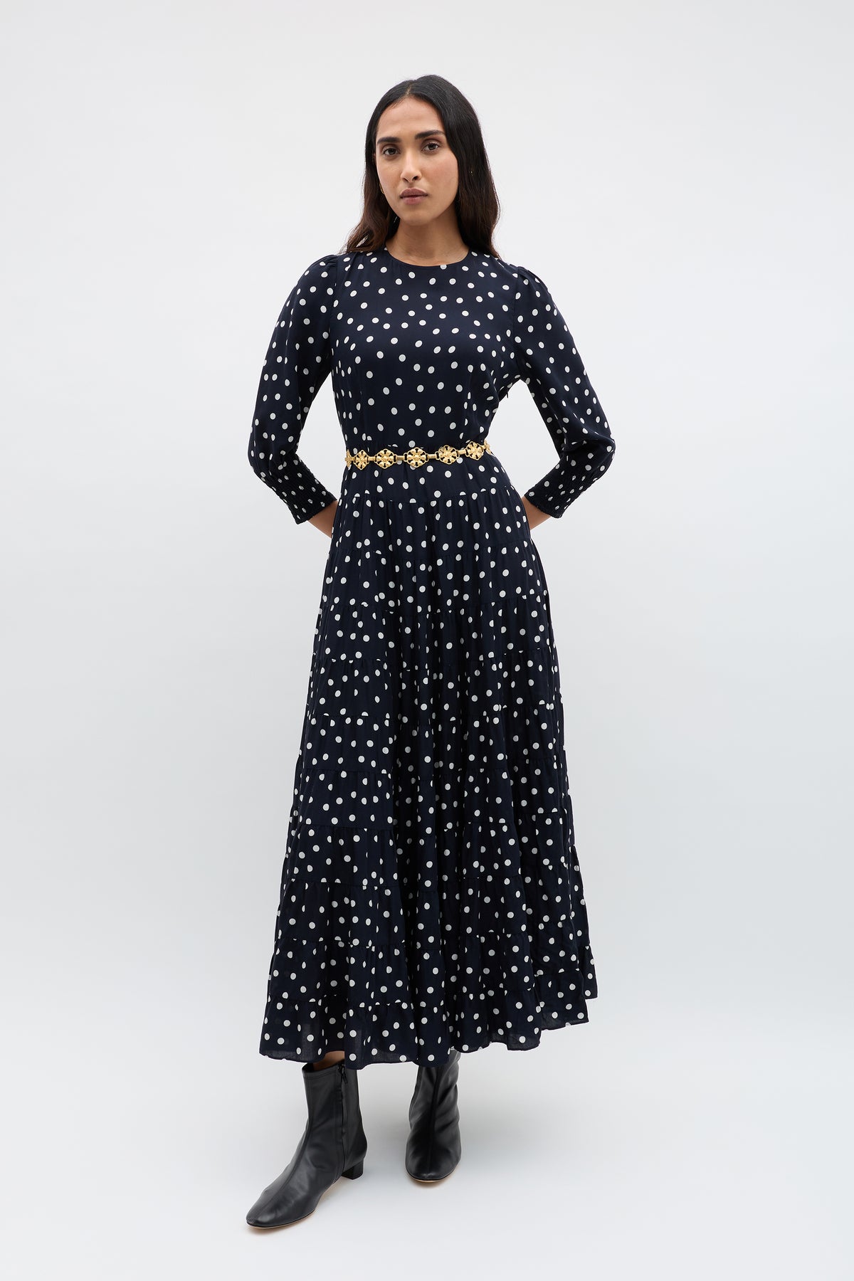 A person stands against a plain background wearing the Kristen - Bohemia Spot Navy Dress, which has long sleeves, a navy spot print, a gold belt at the waist, and black ankle boots.