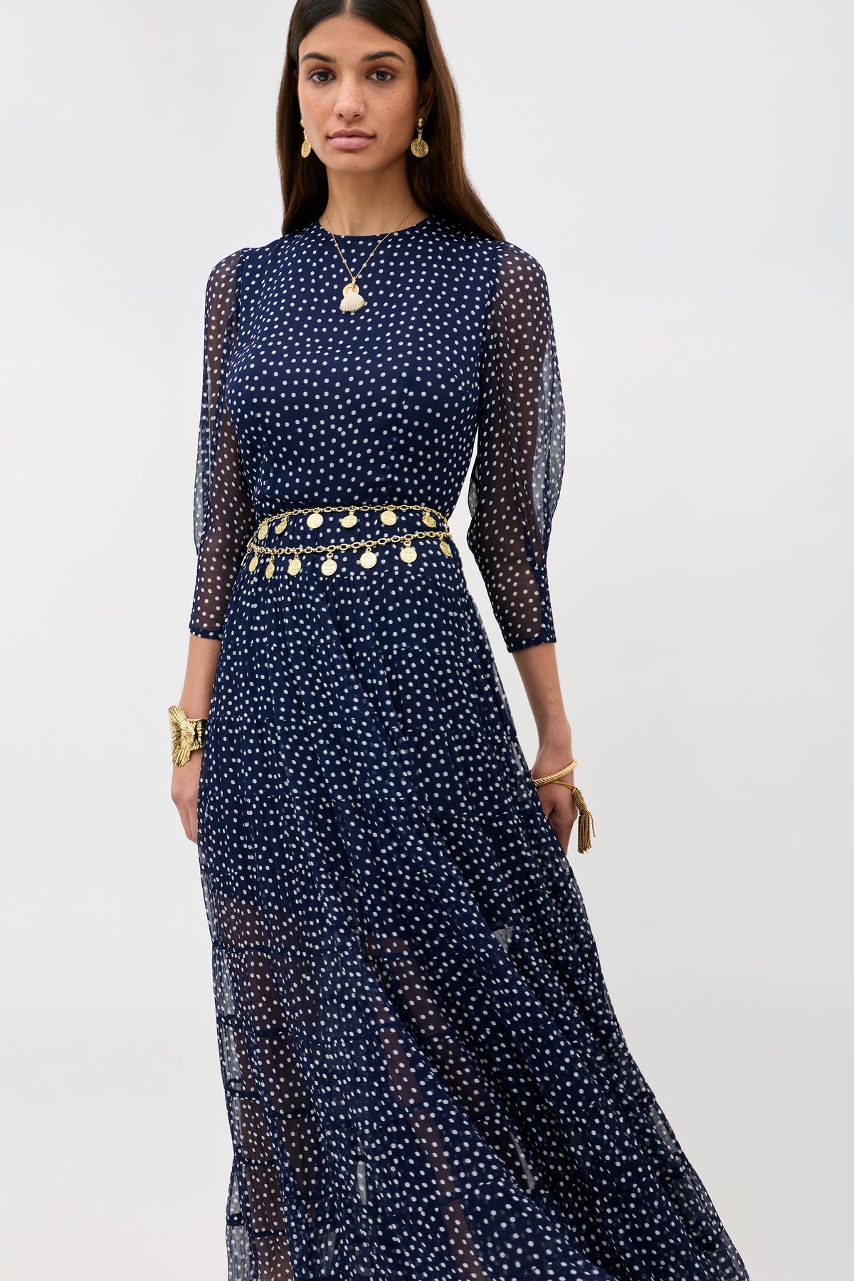 A woman wears the Kristen - Navy Polka Dot georgette dress with ivory spots, tiered skirt, sheer sleeves, and a gold coin belt. She completes her look with gold jewelry and poses against a plain light background.