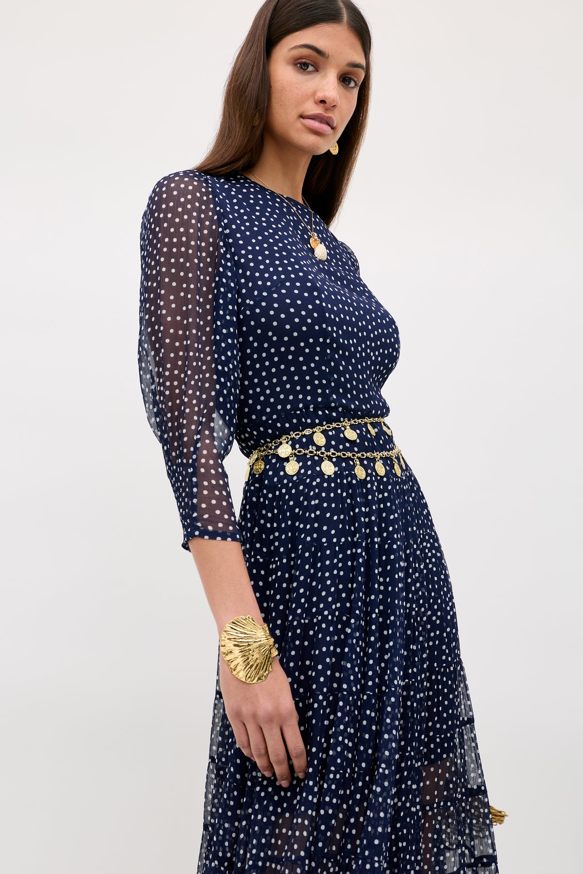 A woman models the Kristen - Navy Polka Dot dress with a tiered skirt, gold coin belt, bracelet, and necklace, standing confidently against a light background.