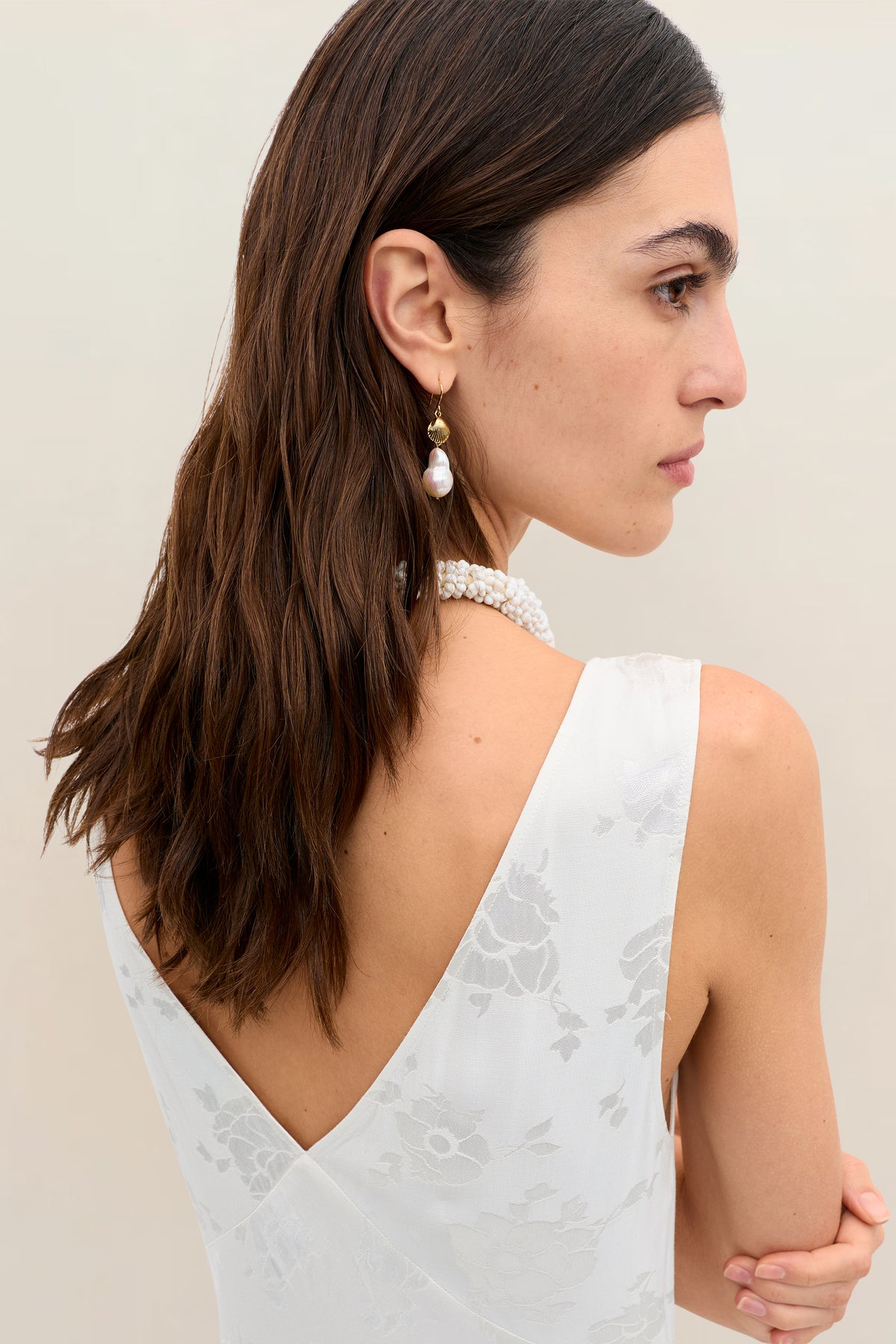 Woman with long brown hair in loose waves wears the Sandrine - Poppy dress: a vintage-inspired, white floral, bias-cut sleeveless design with a deep V back. Styled with a pearl necklace and dangling earring against a light background.