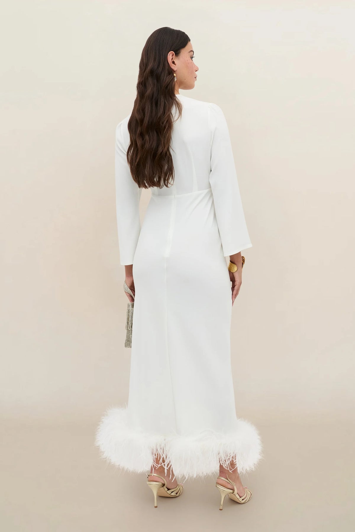 A woman stands facing away, holding a small clutch and wearing the Mya - Ivory Feathers bridal dress—a long-sleeved, bias-cut silk white gown featuring ivory-feather trim at the hem. She has long wavy brown hair and is styled with heeled sandals.