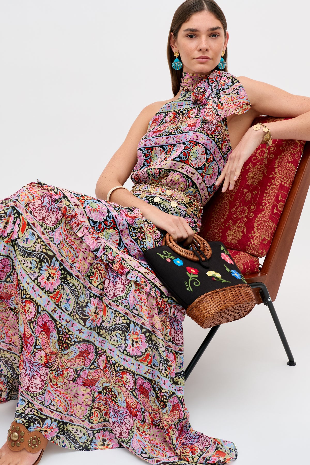 A woman sits on a red patterned chair wearing the Abi - Cara Stripe Black maxi dress with a bow at the neck. She holds a wicker clutch with embroidery and accessorizes with turquoise earrings and gold jewelry.