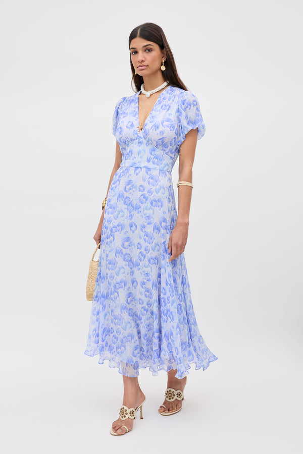 A woman in the Corrine dress—Tulip Fields Cornflower Blue—stands against a plain background, accessorized with gold jewelry, a beaded choker, straw handbag, beige heeled sandals, and wears her straight hair down.