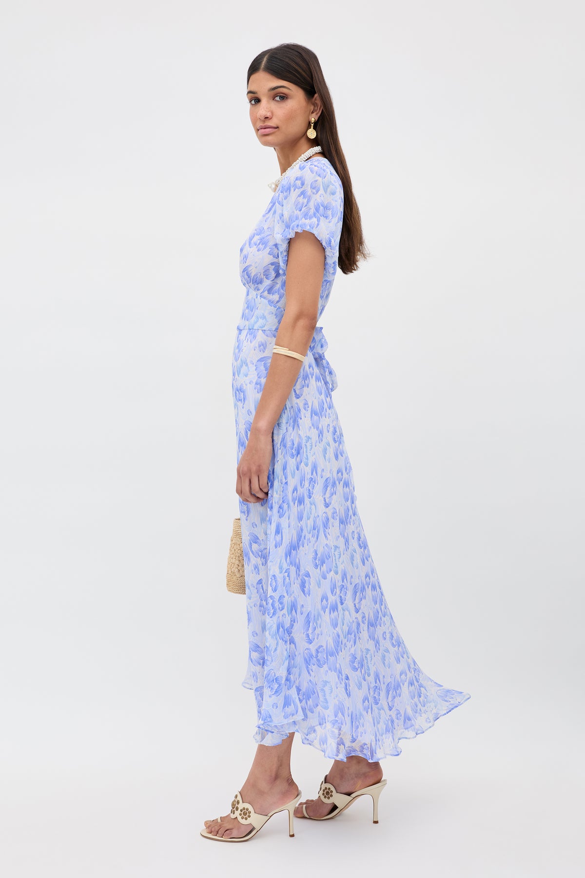 A woman with long dark hair models the Corrine - Tulip Fields Cornflower Blue dress, featuring short blouson sleeves and a flowing hem. She pairs it with gold accessories, beige heeled sandals, and a woven handbag against a white background.