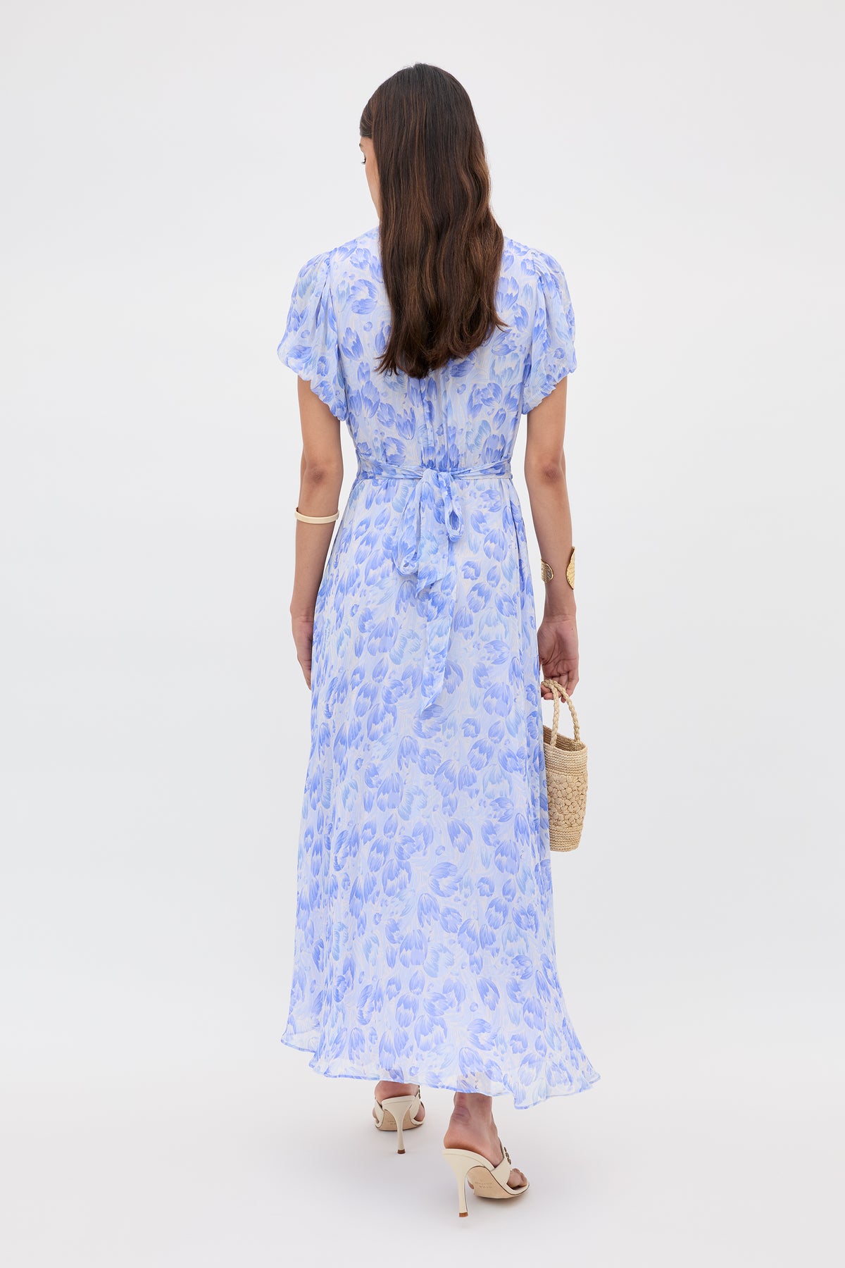 A woman with long brown hair stands facing away, wearing the Corrine dress in Tulip Fields Cornflower Blue, featuring short blouson sleeves. She pairs it with white heeled sandals, a small straw handbag, and a gold bracelet on her left wrist.