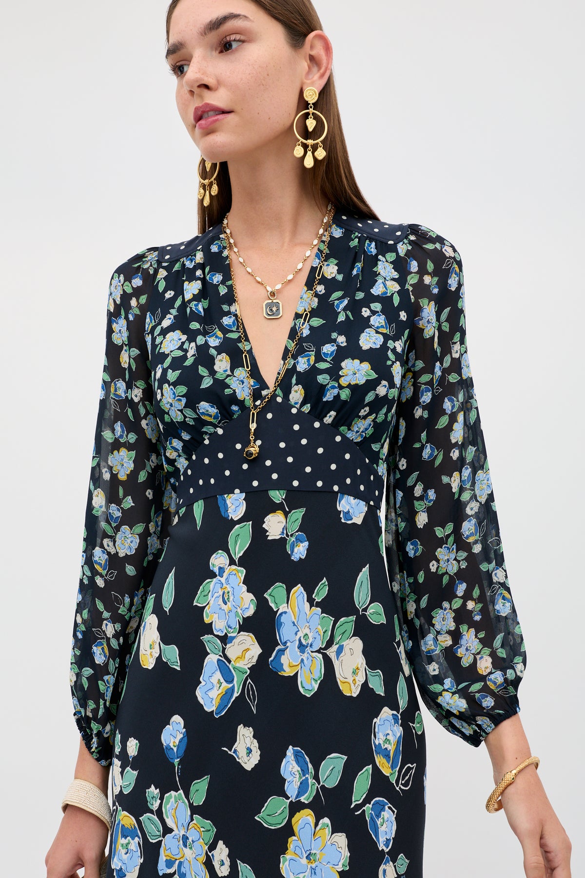 A woman wears the Kaila - Layla Mix Navy dress, featuring blue, green, and yellow floral patterns with white polka dots on the bodice. She completes her look with gold jewelry—layered necklaces and statement earrings.