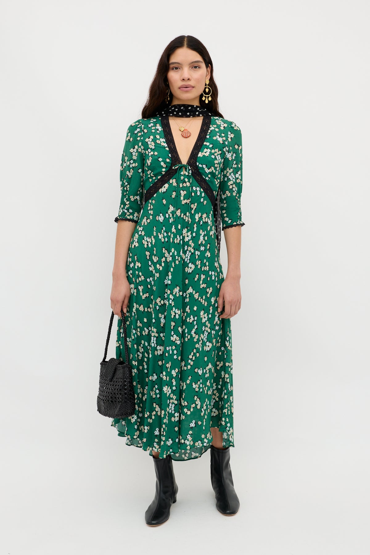 A woman poses against a white background in the Amina - City Floral Olive dress, styled with black lace-trim details, black boots, a neck scarf, gold earrings, and a black woven handbag for a vintage tea dress look.