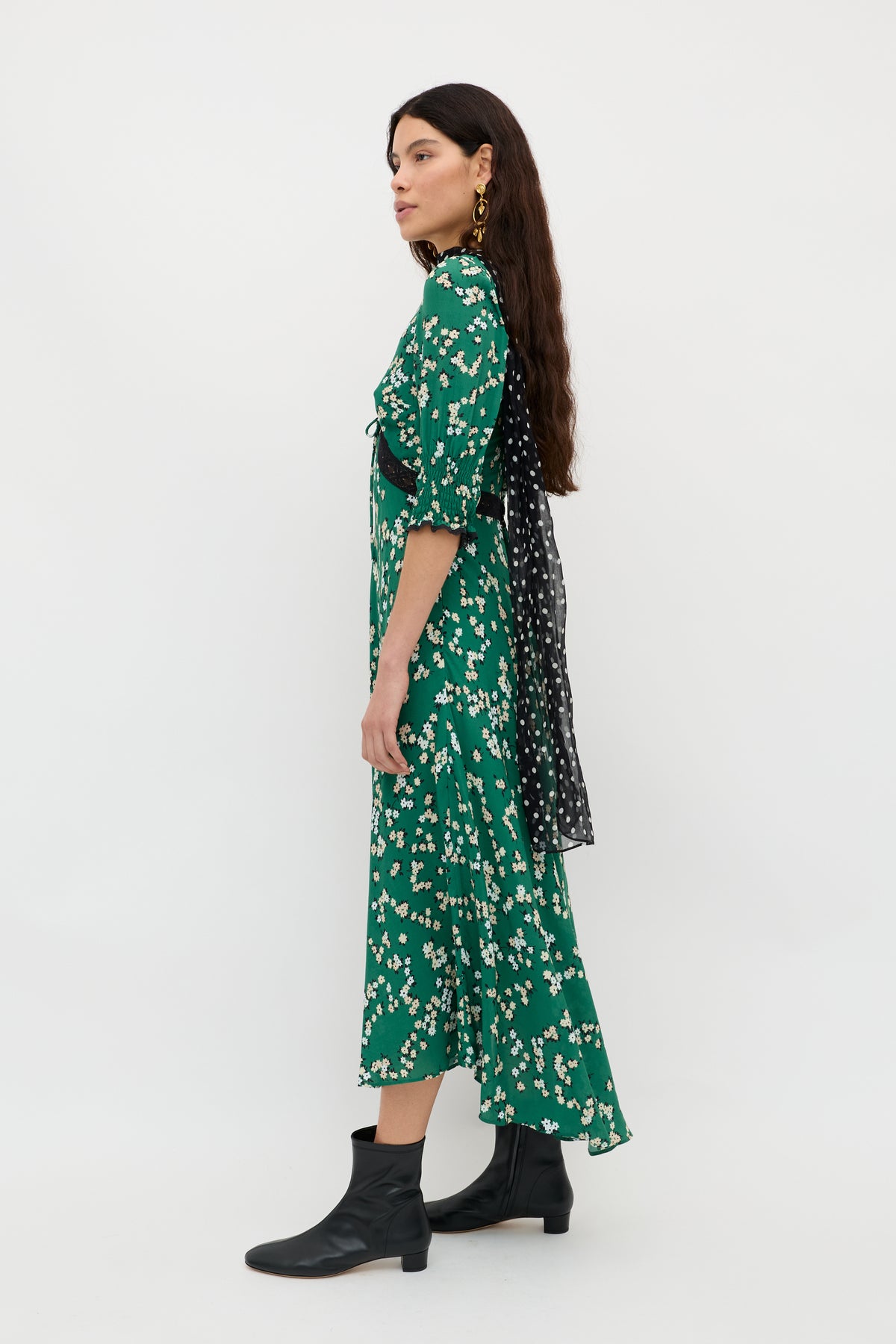 A woman with long dark hair stands in profile against a white background, wearing the "Amina - City Floral Olive" dress styled with a polka dot sash and black ankle boots.