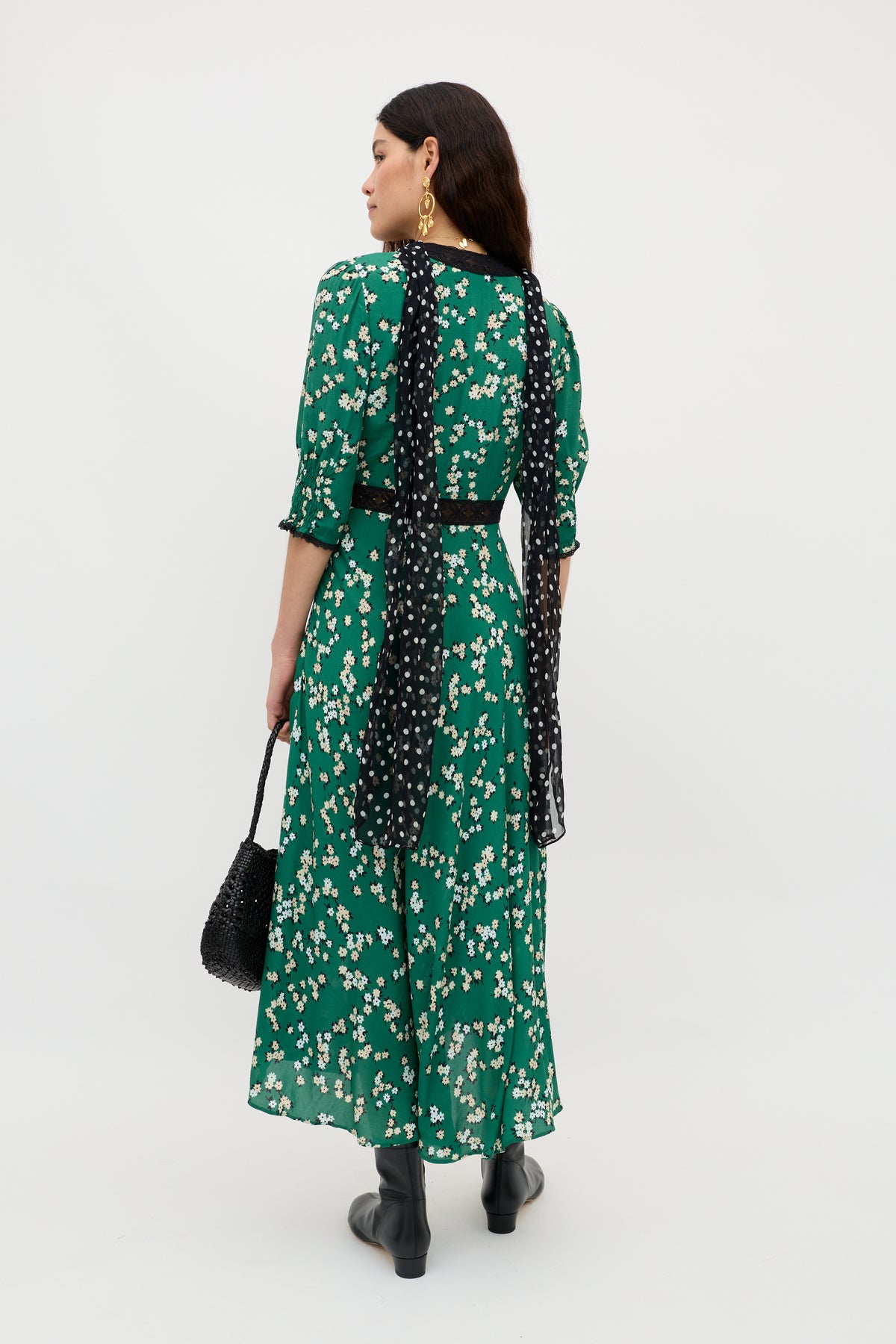 A woman, seen from behind, wears the Amina - City Floral Olive dress with delicate lace trims, paired with black ankle boots, a long black scarf with white dots, gold earrings, and a black textured handbag against a white backdrop.