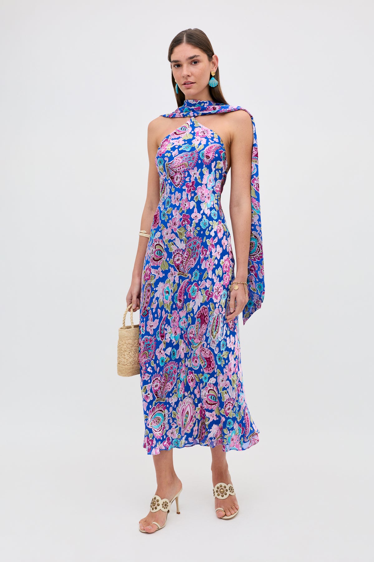 A woman wearing the Hayley - Cara Floral Devore Blue dress with a matching scarf, holding a woven handbag, heeled sandals, and turquoise earrings, stands against a plain white background.
