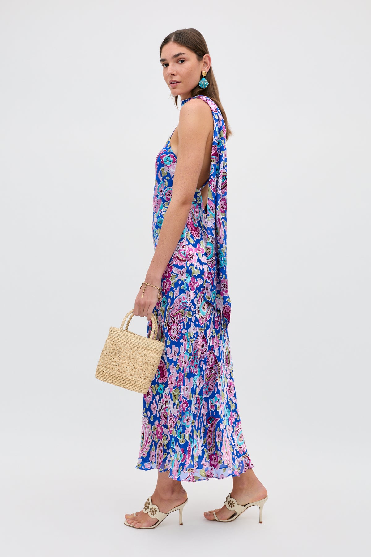 Wearing the “Hayley - Cara Floral Devore Blue” dress, a woman stands against a plain white background. She completes her look with cream heeled sandals, turquoise earrings, and carries a woven straw handbag.