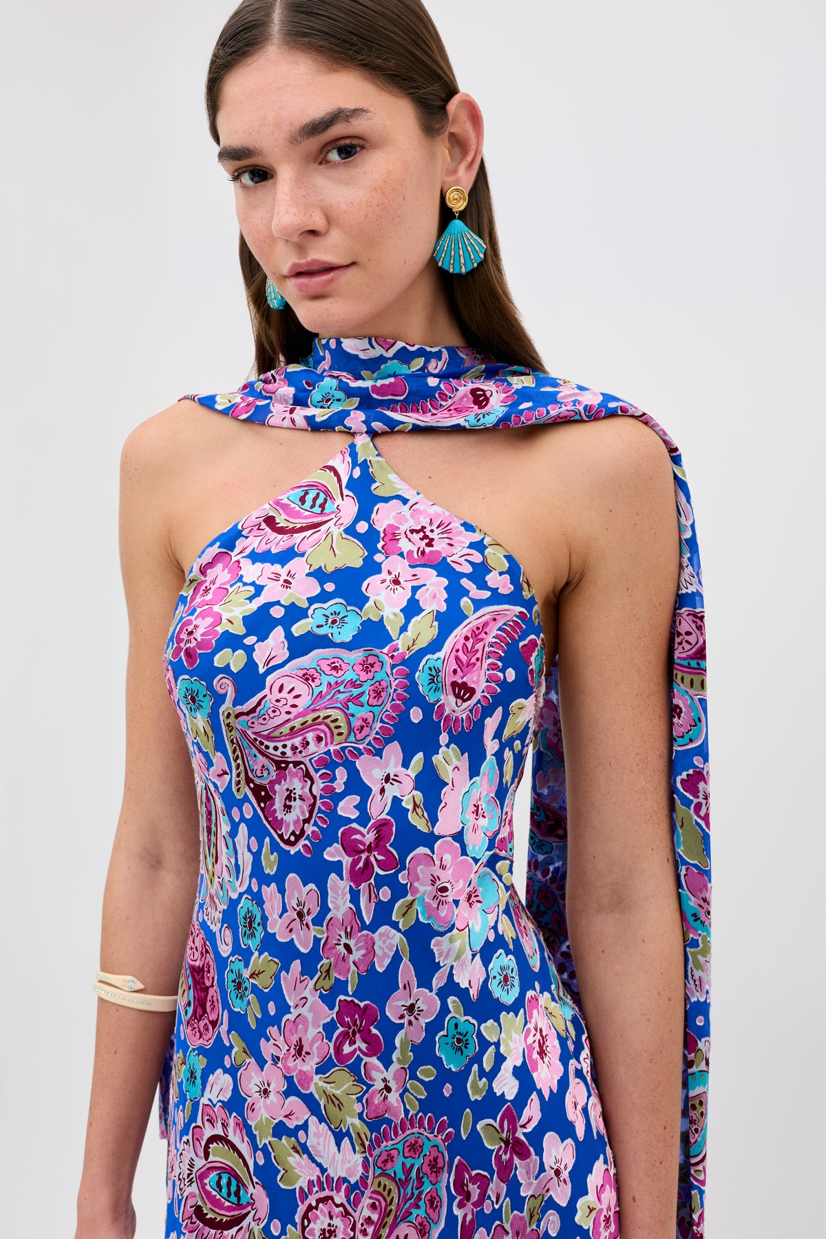 Wearing the Hayley - Cara Floral Devore Blue dress—a sleeveless blue piece adorned with a vibrant floral and paisley pattern—a woman stands against a plain light background, styled with bold tassel earrings and a matching shoulder scarf.