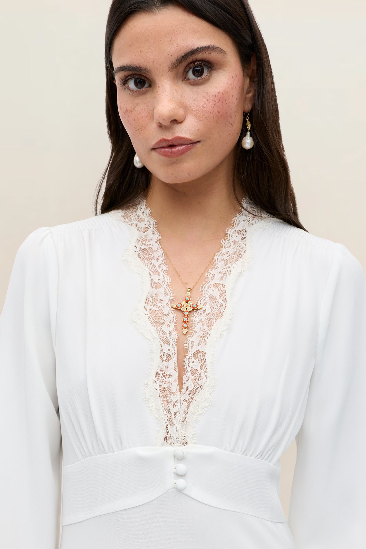 A woman with long dark hair wears the Emory - Ivory Lace gown, featuring satin back crepe details, paired with pearl earrings and a gold cross necklace, looking directly at the camera against a neutral background.