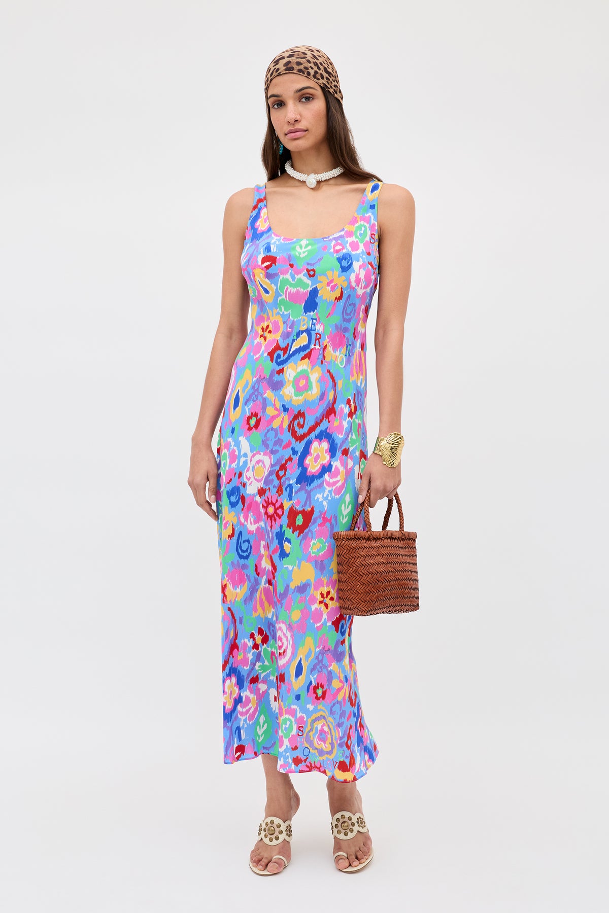 A woman wears the Bondi - Ikat Bloom Lilac maxi dress, a leopard-print headscarf, shell necklace, gold bracelet, woven handbag, and beige sandals with decorative details against a plain white background.