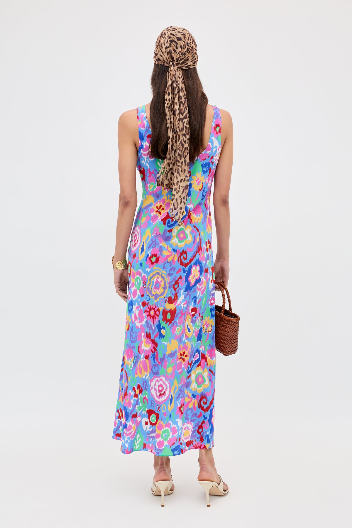 Woman stands with back to camera in the Bondi - Ikat Bloom Lilac dress, beige heeled sandals, leopard print headscarf, and brown woven handbag against a plain light background.