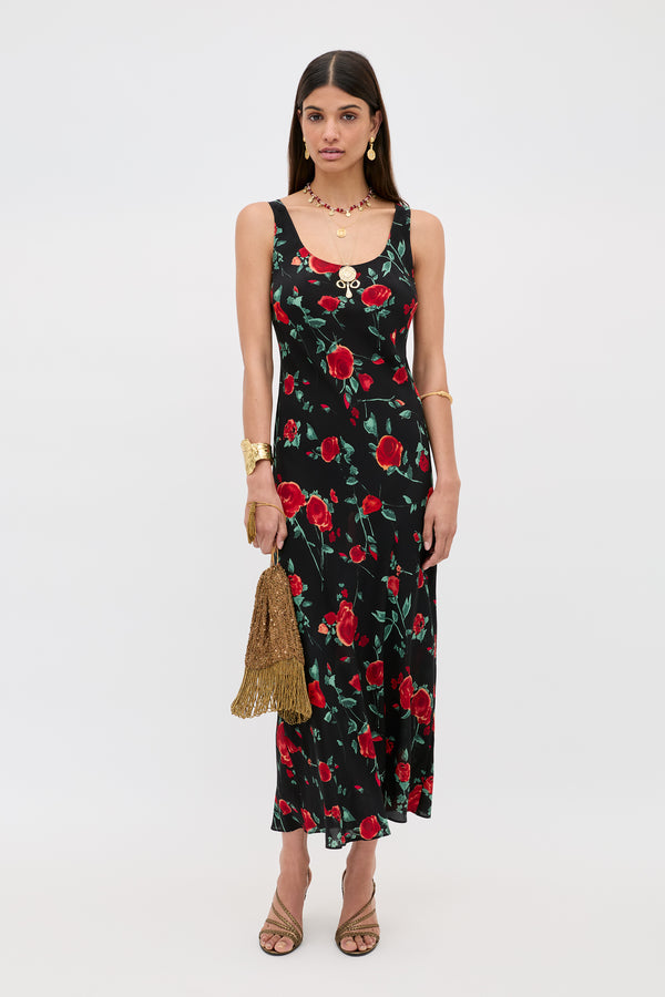 A woman wears the Bondi - Claudia Rose Black, a sleeveless ankle-length black slip dress with a red rose floral print, styled with gold jewelry, nude heels, and a gold tasseled handbag against a plain white background.