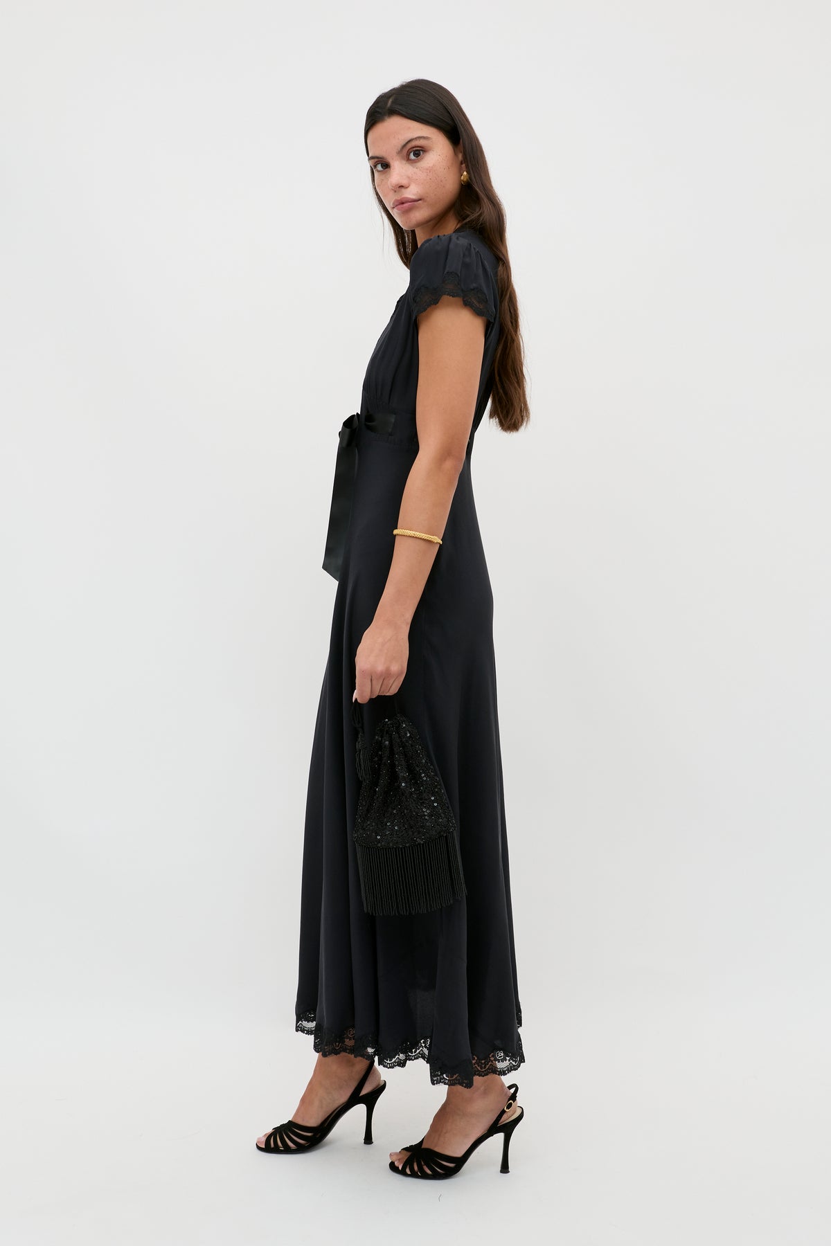 A woman with long brown hair wears the Marlice - Black ankle-length dress with lace trim and a satin bow, styled with black heeled sandals and a matching handbag. She stands sideways against a plain white background.