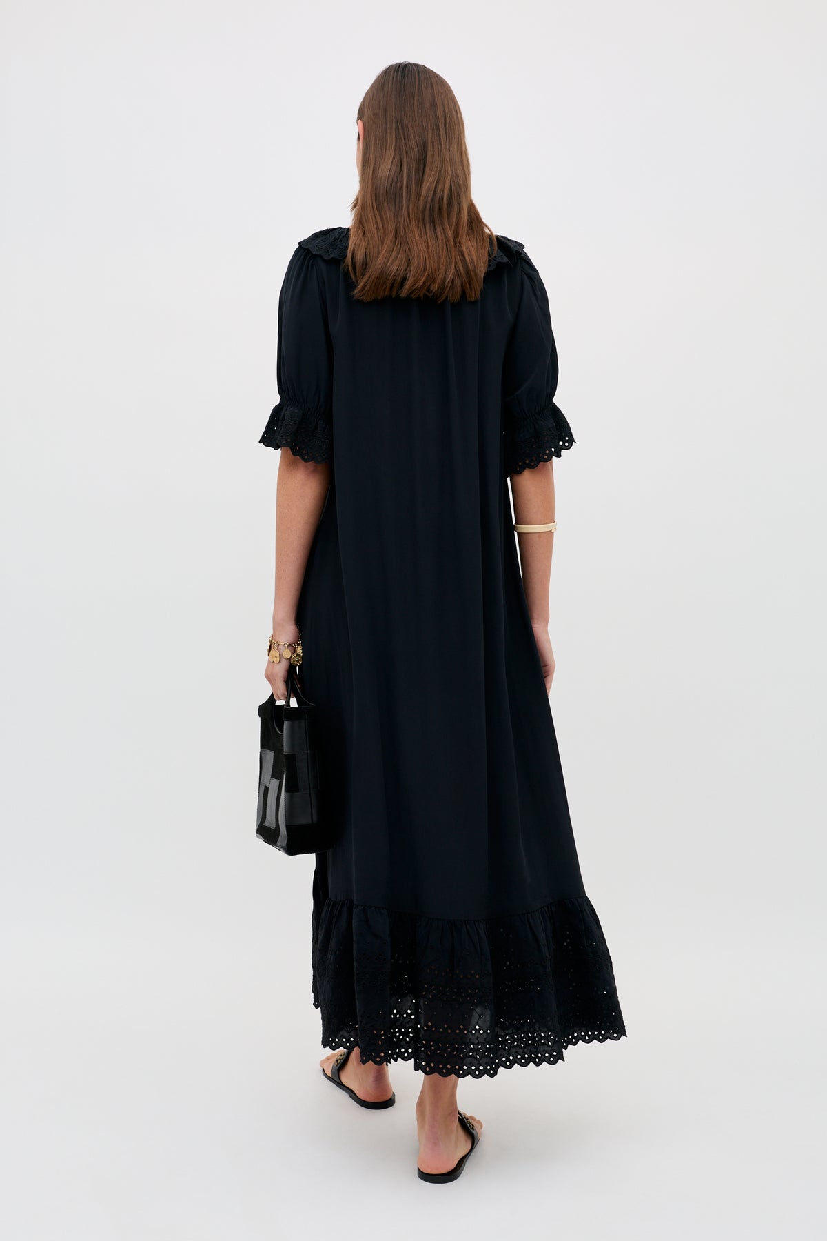 A woman with long brown hair stands facing away, wearing the Aprilia - Gracie Embroidery Black slip dress with lace trim. She holds a black handbag and wears black sandals against a plain, light-colored background.